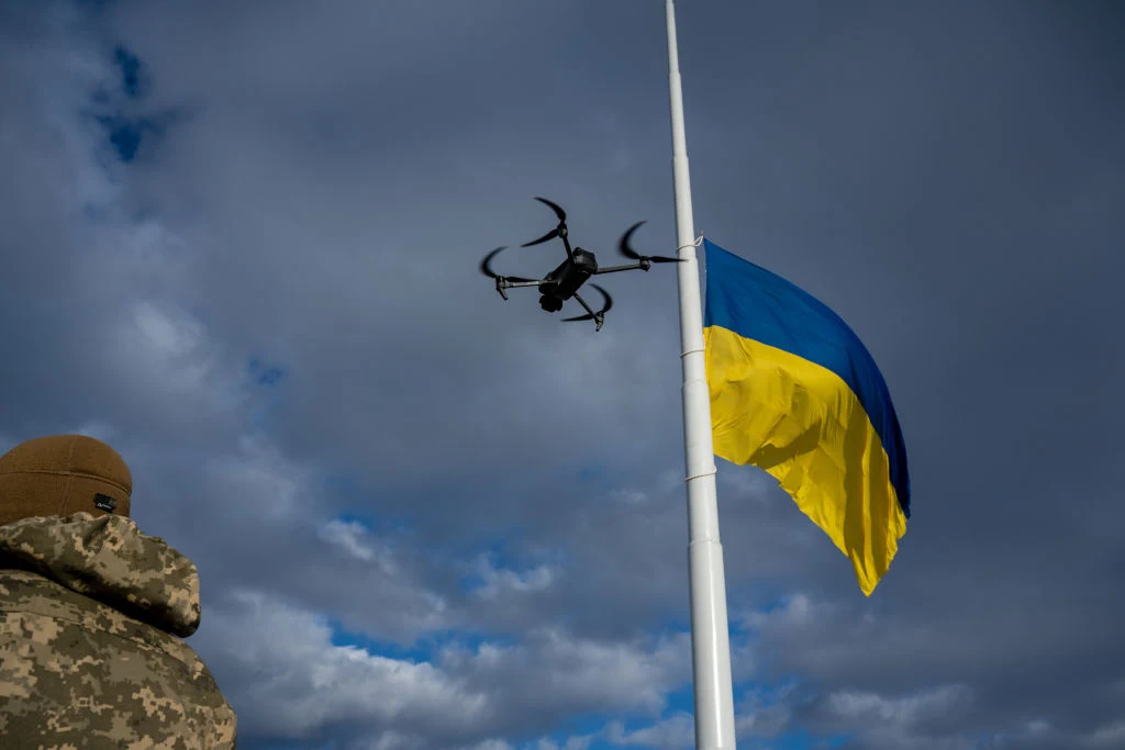 Flaga Ukrainy na maszcie, powiewająca na tle pochmurnego nieba, w pobliżu żołnierz w wojskowym ubraniu oraz unoszący się dron.