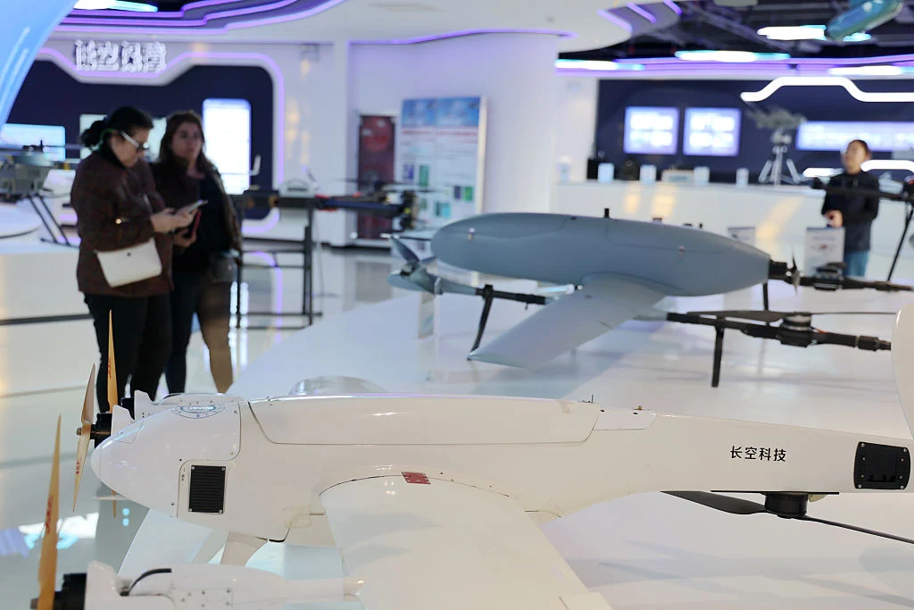 Nowoczesne drony prezentowane w jasnym, futurystycznym wnętrzu wystawienniczym, po lewej stronie trzy osoby oglądające ekspozycję i robiące notatki, w głębi widoczne interaktywne ekrany i inne technologie.