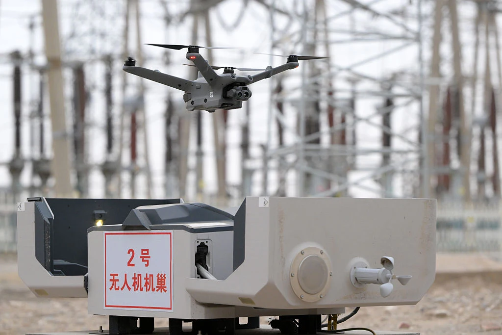 Dron startujący z platformy dokującej na tle infrastruktury energetycznej, otoczony elementami przemysłowymi i przewodami.