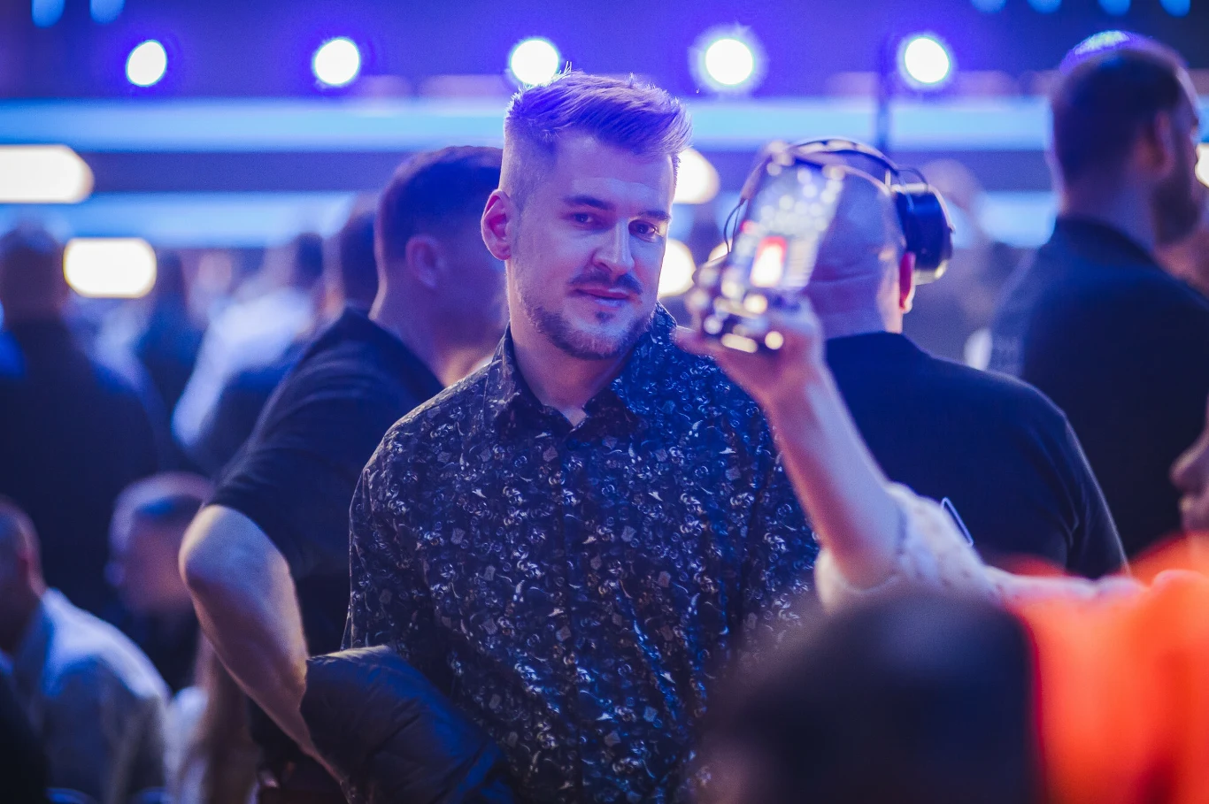 Ujęcie młodego mężczyzny stojącego w tłumie ludzi podczas wydarzenia rozrywkowego, dynamiczne światła w tle oraz osoby w ruchu, jedna z postaci trzyma przezroczysty kubek.