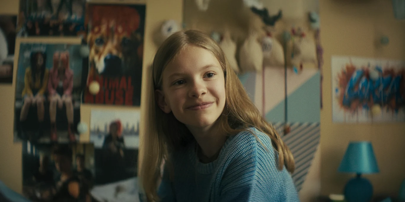 "Psoty" Uśmiechnięta dziewczynka siedzi w pokoju z jasnoniebieską lampką na biurku i kolorowymi plakatami na ścianach, w tle pluszowe zabawki zawieszone nad łóżkiem.