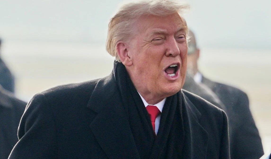 Mężczyzna w średnim wieku w eleganckim, czarnym płaszczu oraz czerwonym krawacie, stojący na zewnątrz, z otwartymi ustami jakby w trakcie wypowiedzi lub okrzyku. W tle rozmyte sylwetki postaci i nieostre otoczenie.