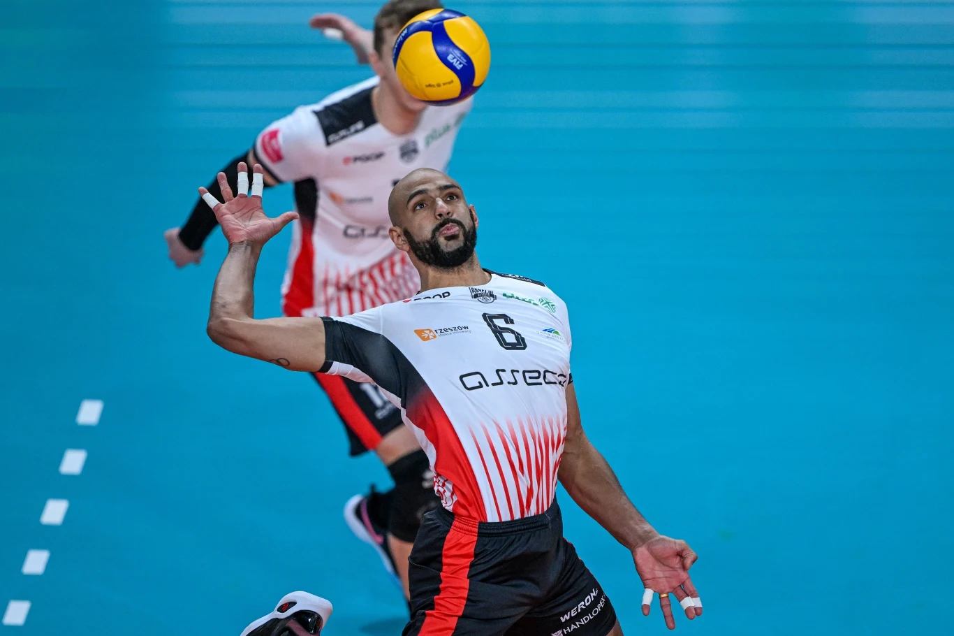 Zawodnik siatkarski w biało-czerwono-czarnym stroju podczas dynamicznej akcji z piłką unoszącą się nad jego głową, w tle inny zawodnik na niebieskim boisku do siatkówki.