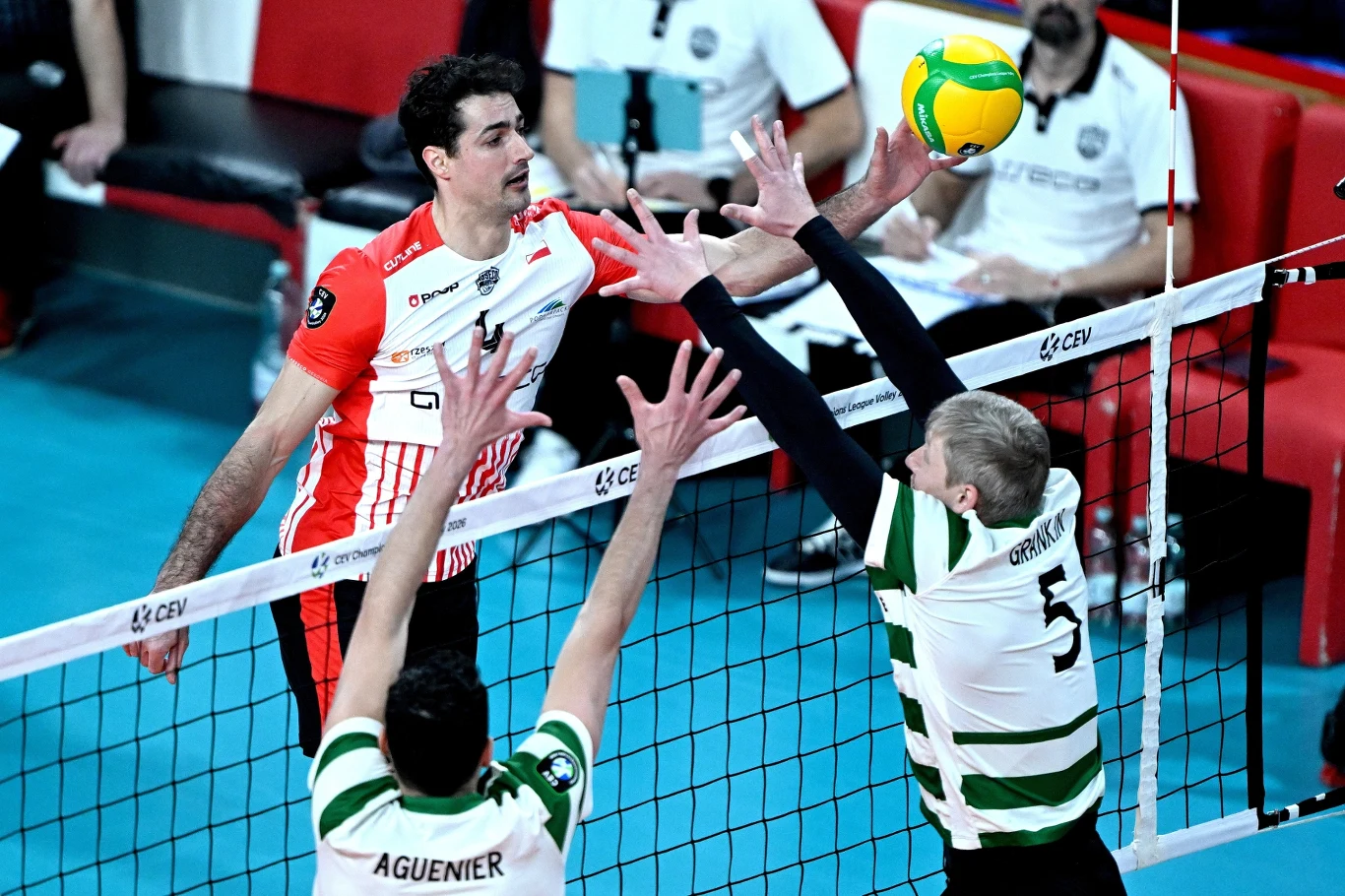 Zawodnik w czerwono-białym stroju wykonuje atak po lewej stronie siatki, próbując przebić piłkę ponad dwoma blokującymi w biało-zielonych strojach. Piłka znajduje się tuż nad siatką. W tle widoczni sędziowie i ławka rezerwowych.