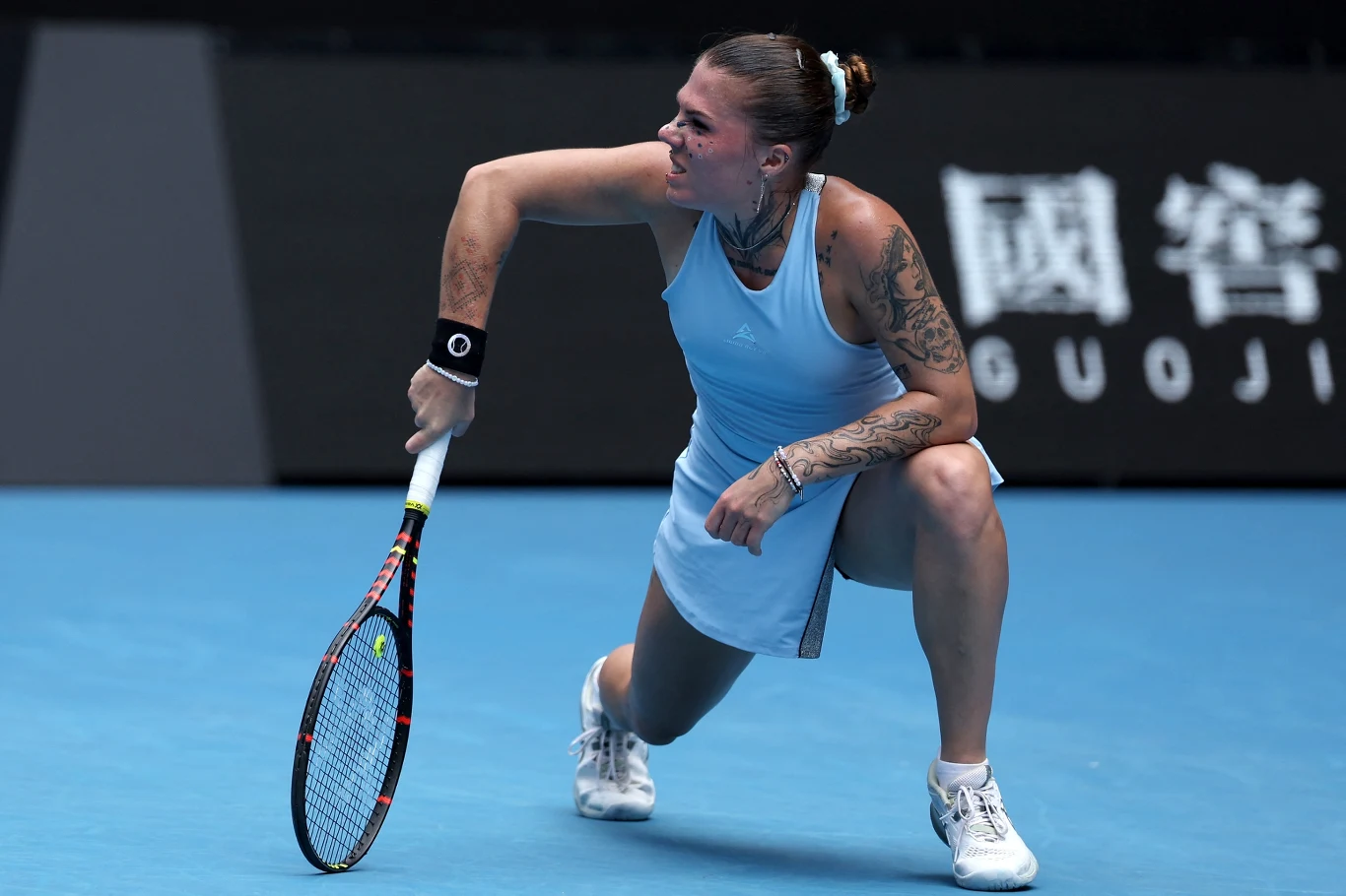 Tenisistka podczas dynamicznego uderzenia piłki na korcie, w widocznym skupieniu, ubrana w jasnoniebieską sukienkę sportową, z tatuażami na rękach i widoczną rakietą tenisową.