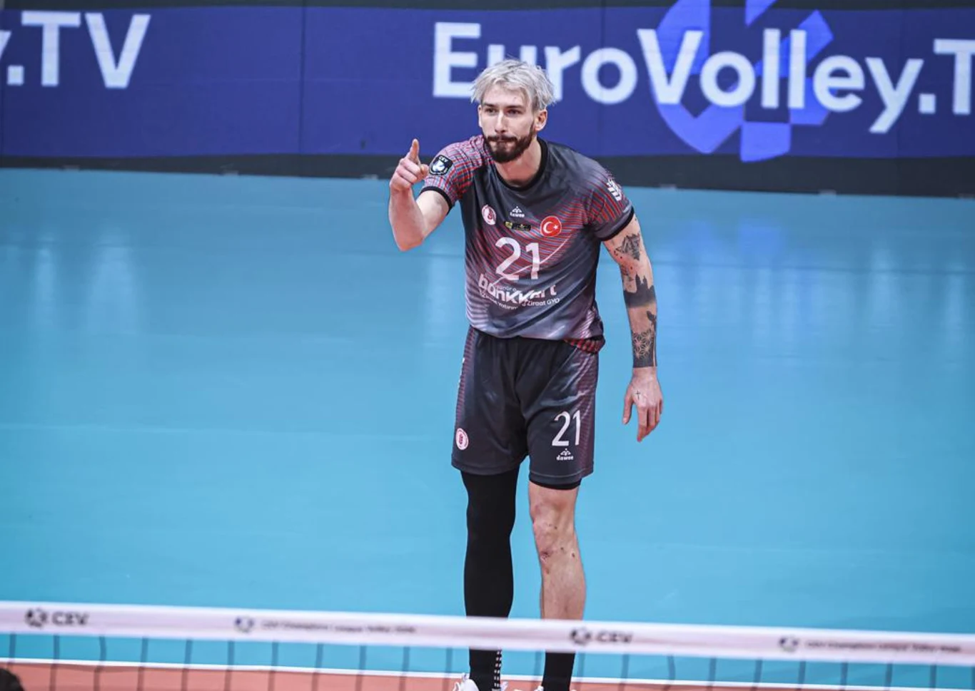 Zawodnik siatkarski w ciemnoszarym stroju reprezentacji Turcji, z numerem 21, stoi na boisku przy siatce, wykonując gest dłonią. W tle widoczny fragment tablicy reklamowej EuroVolley.