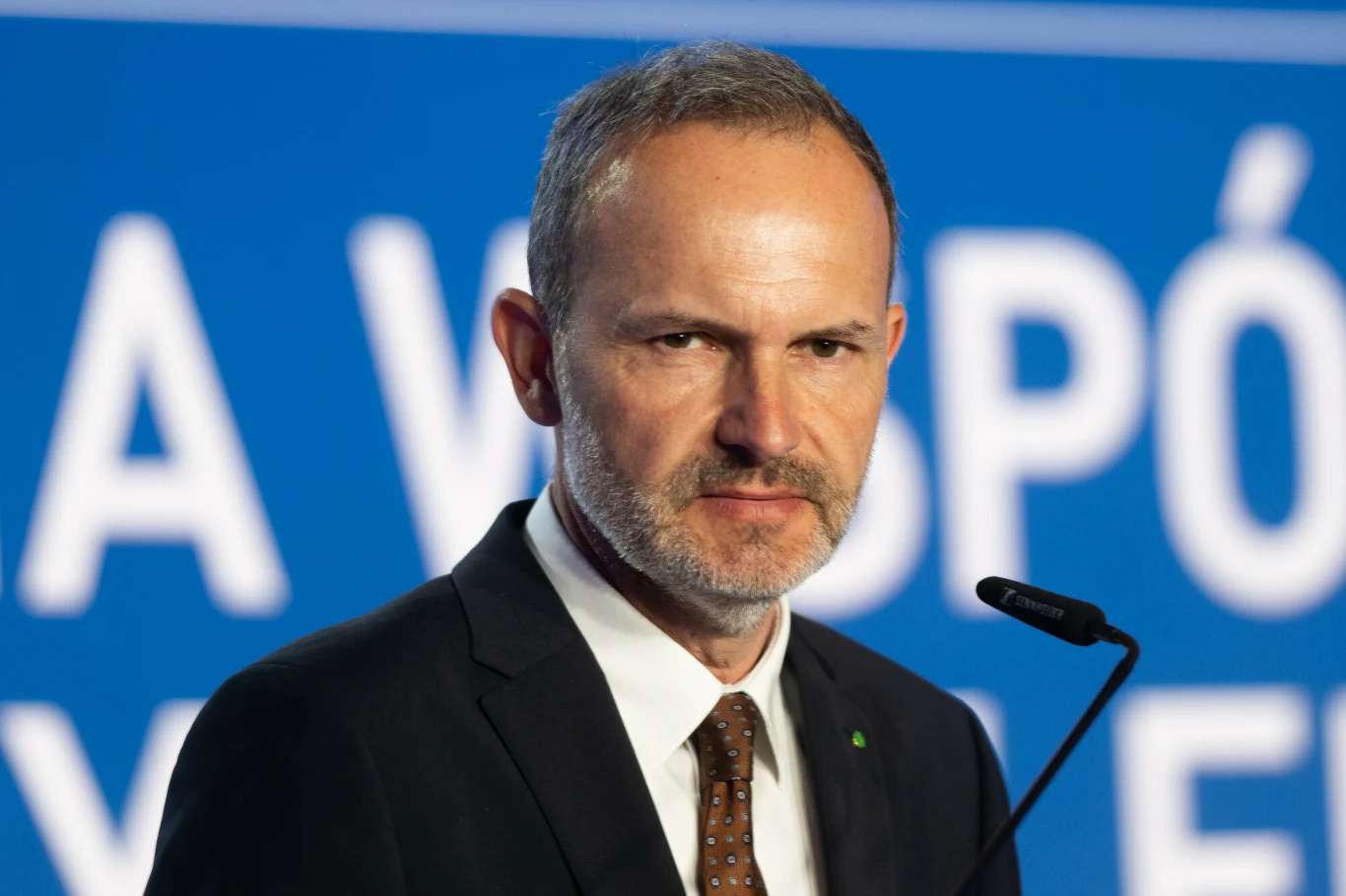 Mężczyzna w garniturze i krawacie stoi przy mikrofonie na tle niebieskiej tablicy z białym napisem, patrzy poważnie w obiektyw.