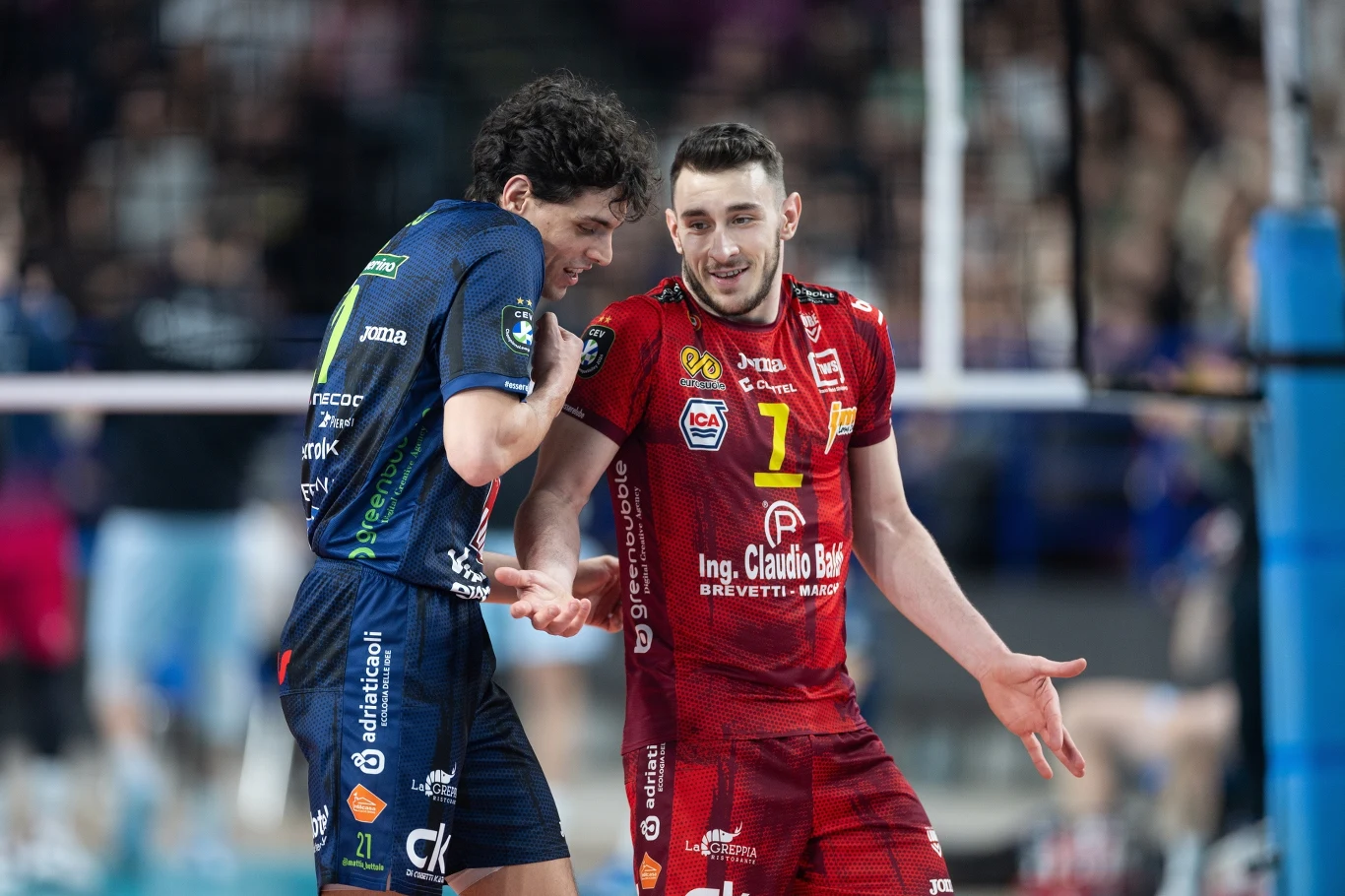 Dwóch siatkarzy w trakcie rywalizacji sportowej na hali, ubranych w stroje klubowe – jeden w czerwonym stroju, drugi w niebieskim, prowadzących ze sobą konwersację przy siatce, w tle widoczna rozmyta publiczność.