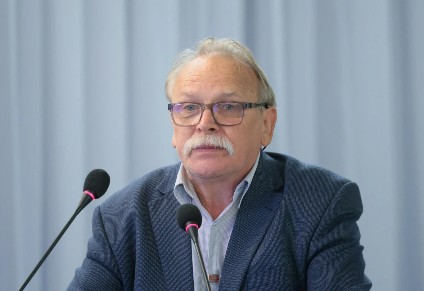 Starszy mężczyzna z wąsem w garniturze i okularach siedzi przy dwóch mikrofonach na tle jasnej kotary, sprawiając wrażenie uczestnika konferencji lub spotkania medialnego.