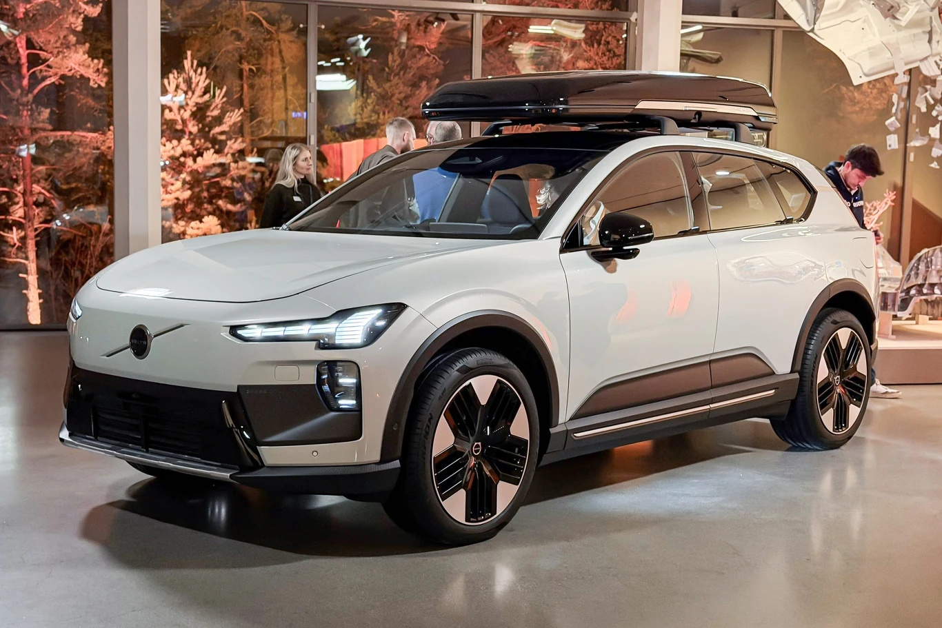 Jasnoszary nowoczesny samochód elektryczny SUV z zamontowanym na dachu boxem bagażowym stoi w oświetlonym wnętrzu, na drugim planie widoczne są rozmyte sylwetki ludzi oraz elementy wystroju z motywem drzew.