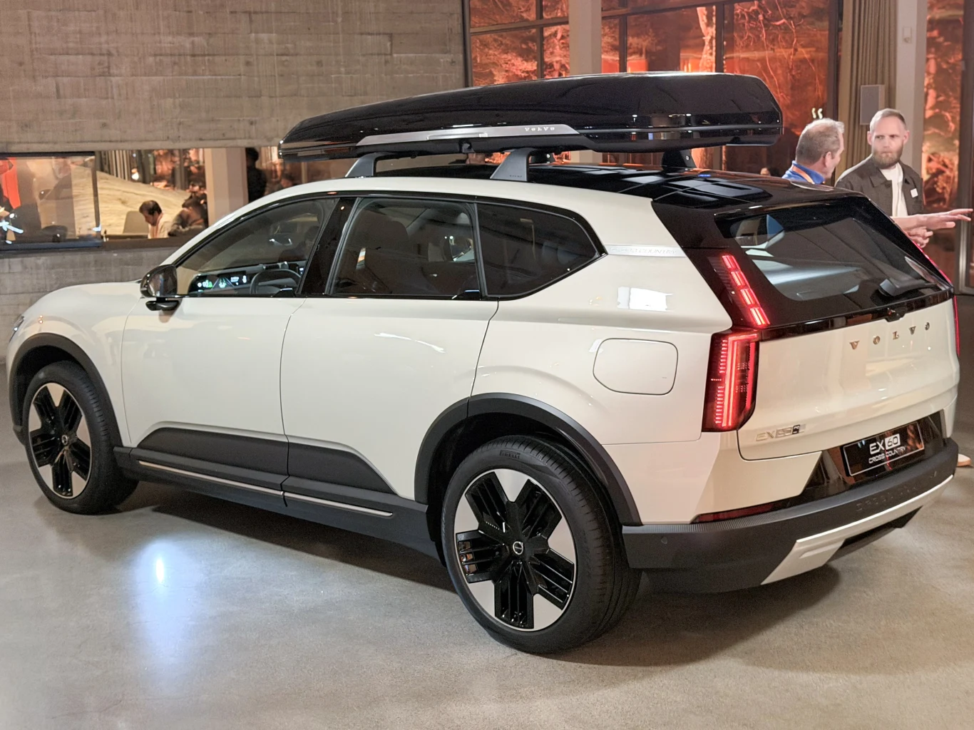 Biały samochód elektryczny marki Volvo z czarnym boksem dachowym stoi zaparkowany w pomieszczeniu o nowoczesnym wystroju. W tle widać dwie osoby rozmawiające oraz duże okno z widokiem na oświetloną ulicę.