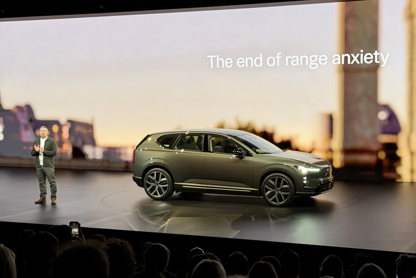 Nowoczesny samochód elektryczny prezentowany na scenie podczas konferencji, z napisem podkreślającym koniec obaw związanych z zasięgiem, obok prowadzącego prezentację.
