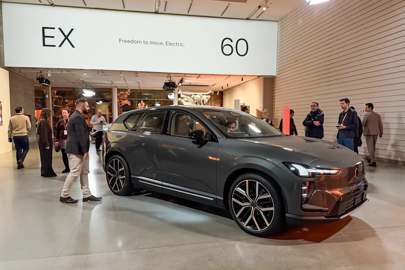 Nowoczesny samochód elektryczny w ciemnoszarym kolorze stoi w jasnej przestrzeni ekspozycyjnej, wokół auta zgromadzeni są ludzie, którzy przyglądają się pojazdowi. W tle widoczna jest tablica z napisem EX60, podkreślająca model auta oraz hasło o mobiln...