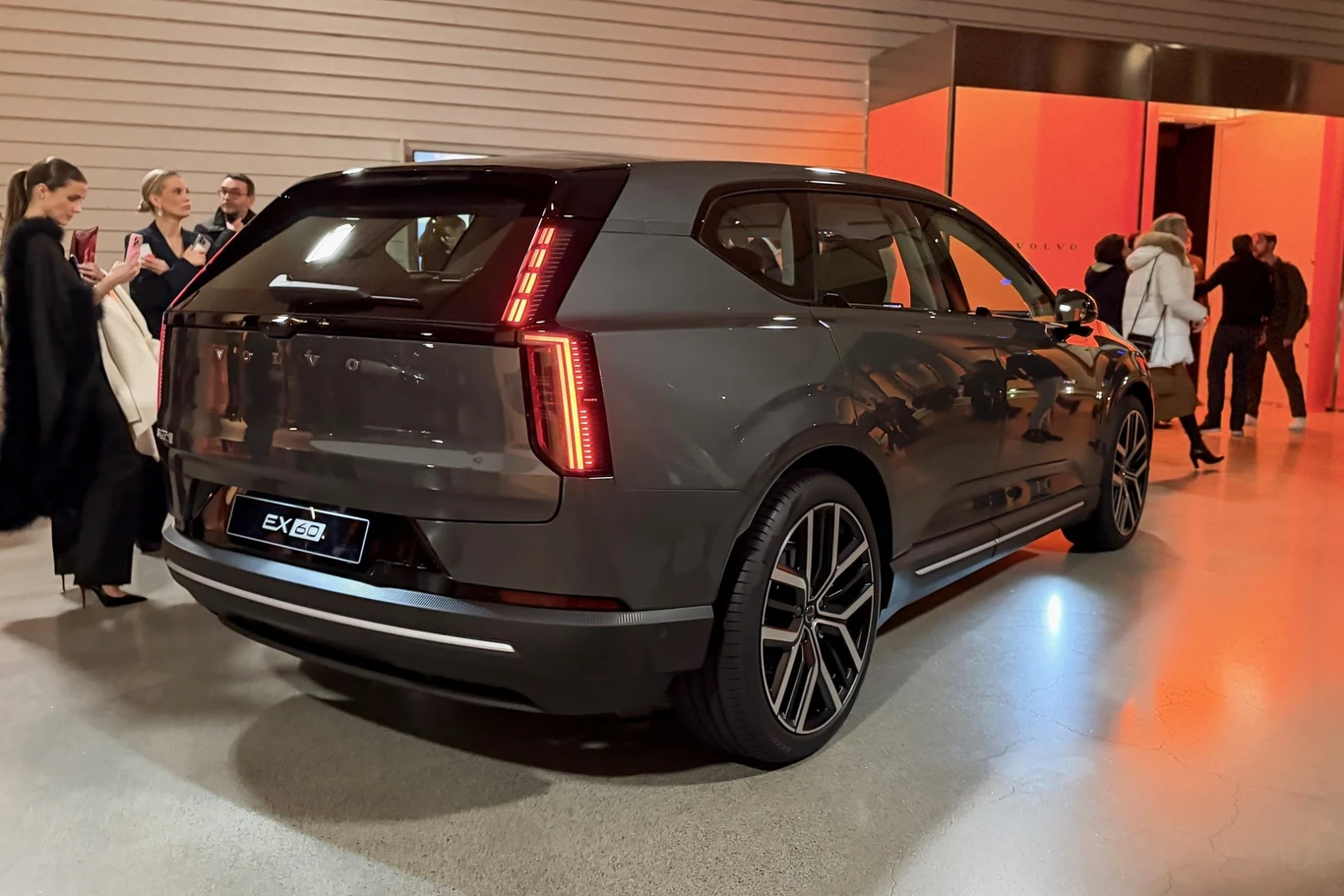 Szary nowoczesny SUV z charakterystycznymi pionowymi światłami tylnymi stoi zaparkowany w przestronnym, dobrze oświetlonym pomieszczeniu. W tle znajduje się grupa elegancko ubranych osób rozmawiających i robiących zdjęcia.