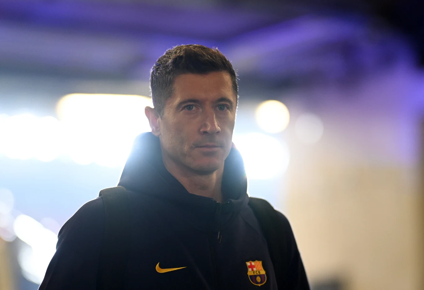 Mężczyzna w sportowej bluzie z logo FC Barcelony, patrzący prosto przed siebie, lekko oświetlony tylnym światłem na rozmytym tle.