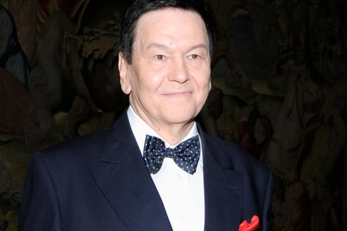 Bogusław Kaczyński Elegancko ubrany mężczyzna w ciemnym garniturze z białą koszulą i muchą w kropki, z czerwonym poszetką, stoi na tle ozdobnej, ciemnej tapiserii.