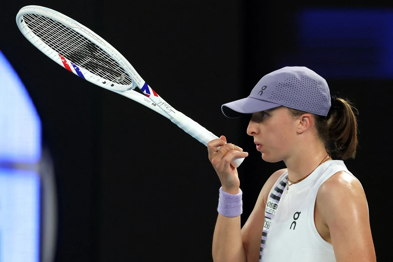 Kobieta w sportowym stroju, skupiona, trzyma w ręku rakietę tenisową podczas meczu na korcie. Na głowie ma fioletową czapkę z daszkiem, na nadgarstku opaskę. Tło rozmyte, uwaga skupiona na zawodniczce i rakiecie.