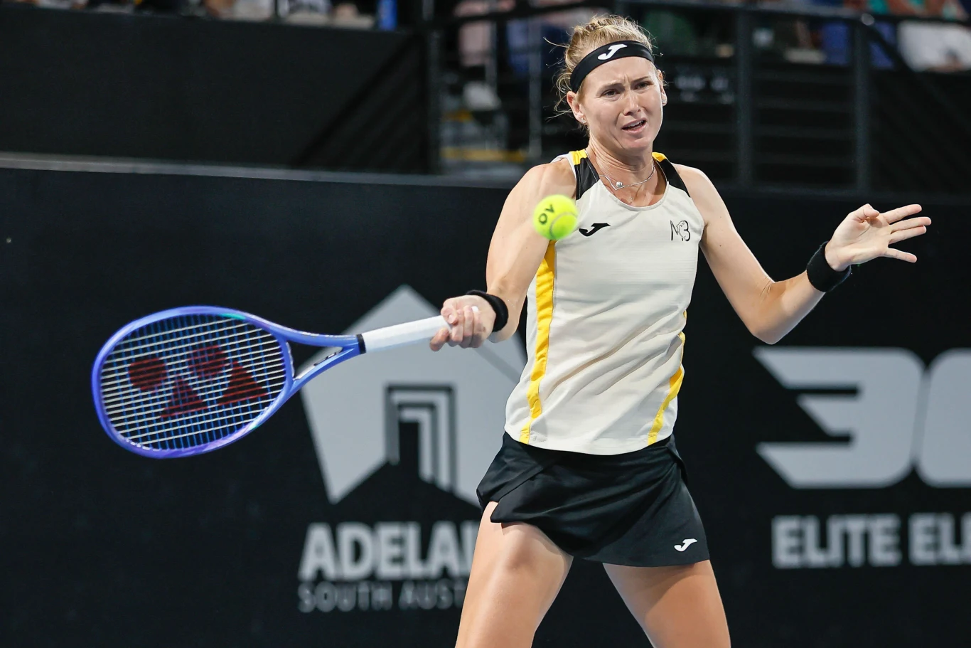 Tenisistka w trakcie wykonywania uderzenia forehandem podczas meczu na korcie, z wyraźnie widoczną rakietą i piłką, w tle widoczne elementy hali sportowej oraz reklamy turnieju.