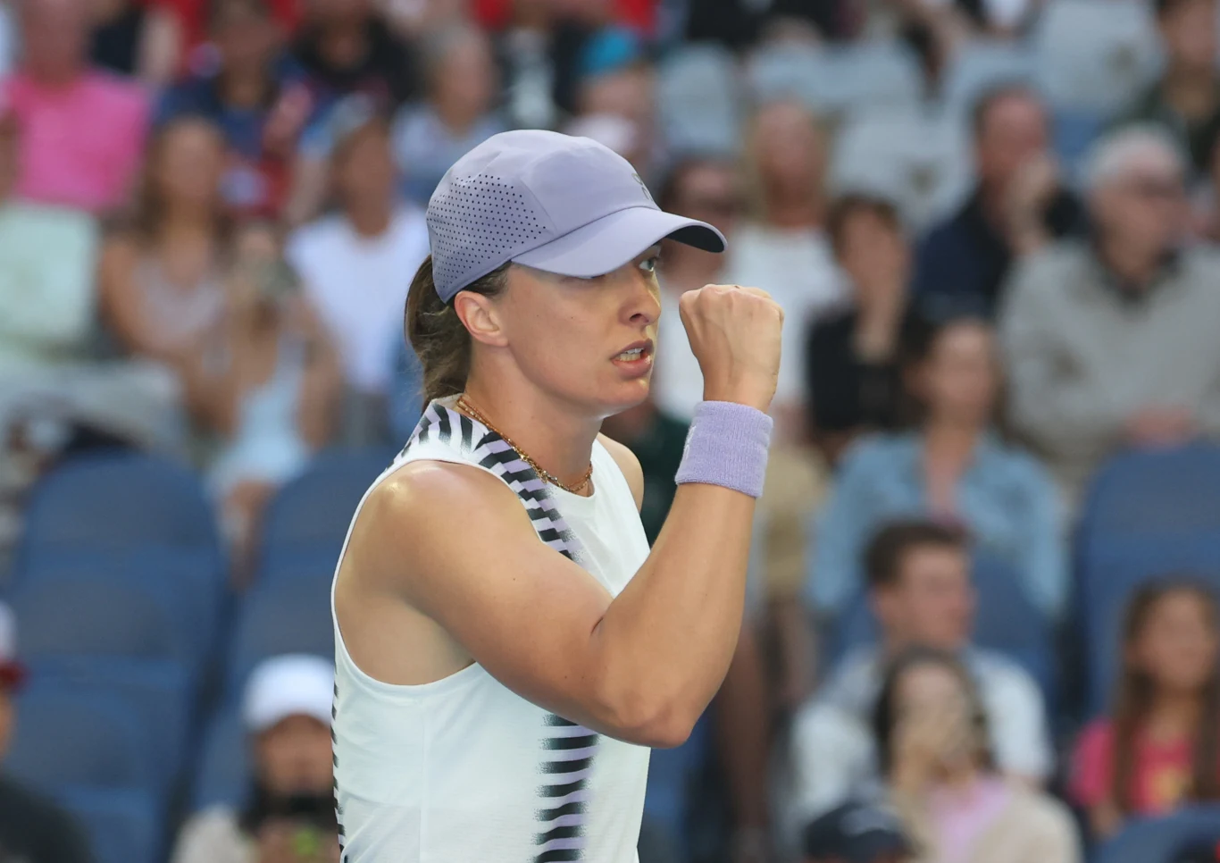 Młoda kobieta w sportowym stroju i czapce z daszkiem wykonuje gest triumfu na tle rozmytego tłumu siedzącego na trybunach.