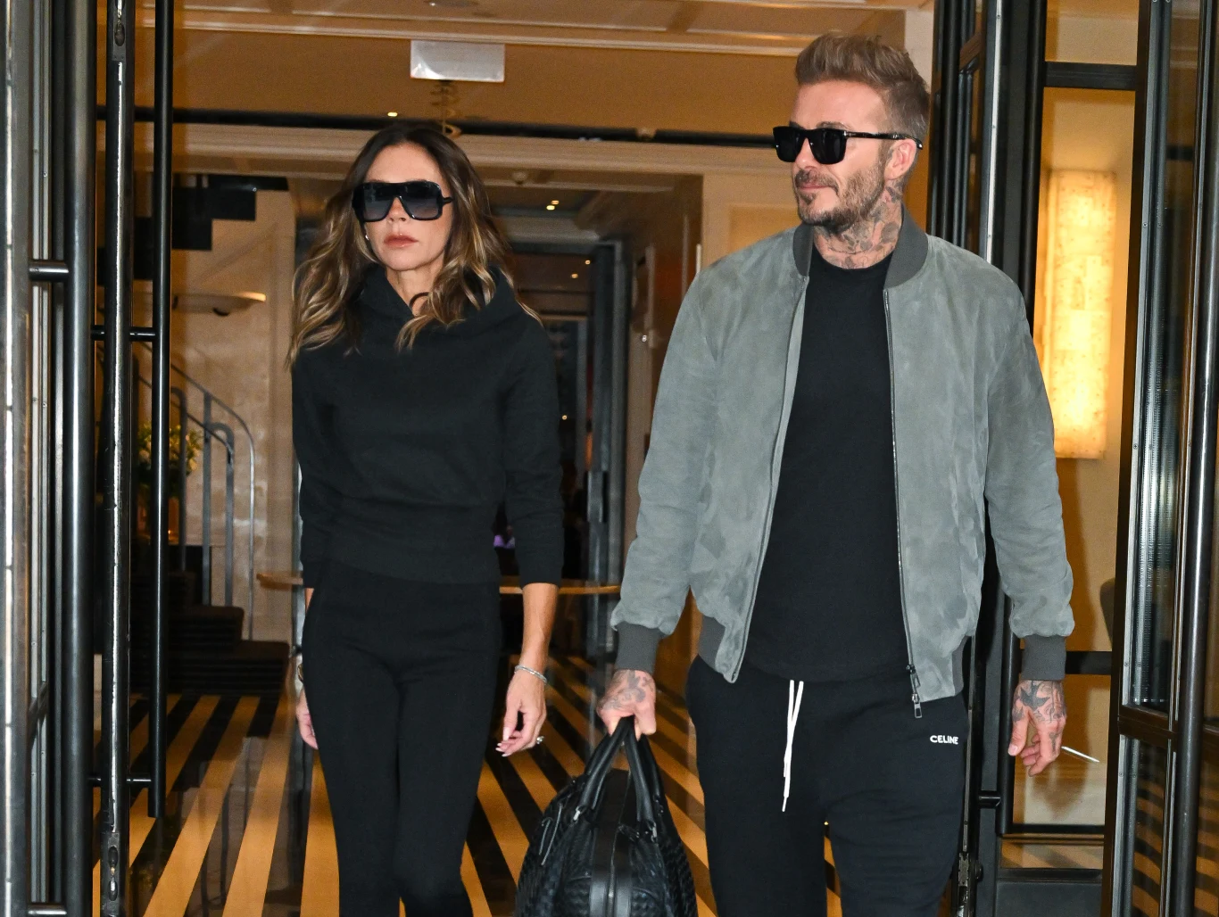 Para ubrana w casualowe stroje, oboje noszą okulary przeciwsłoneczne, wychodzą razem przez szklane drzwi budynku. Mężczyzna trzyma czarną torbę, wnętrze zdobią podłoga w biało-czarne pasy i ciepłe oświetlenie.