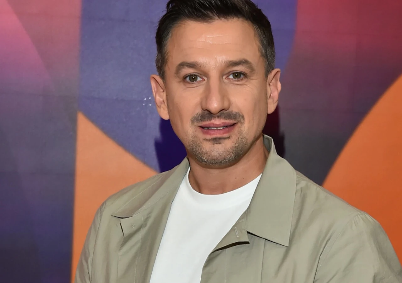 Maciej Rock Uśmiechnięty mężczyzna w jasnej marynarce i białej koszulce stoi przed kolorowym tłem w odcieniach fioletu i pomarańczu. Krótko ostrzyżone włosy i lekki zarost podkreślają zadbany wygląd.