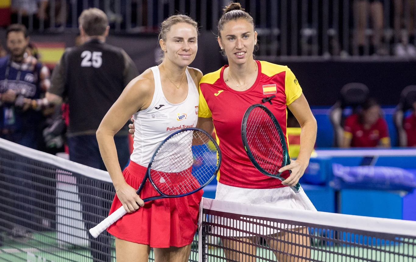 Dwie tenisistki stoją przy siatce na korcie, trzymając rakiety tennisowe, ubrane w stroje sportowe w barwach swoich krajów, otoczone przez trybuny z widownią i sędziów w tle.