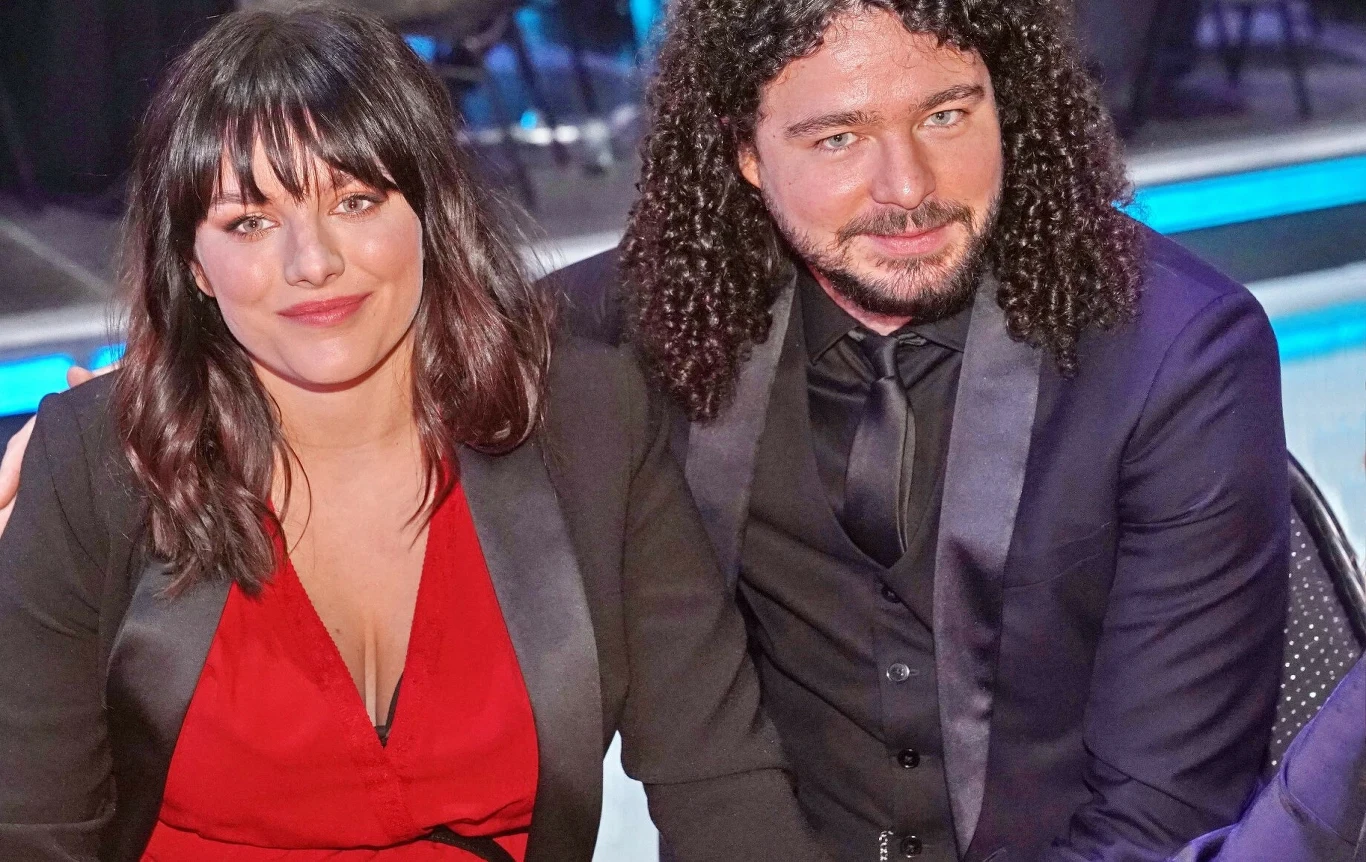 Ewa Farna i Martin Chobot Dwie uśmiechnięte osoby siedzące obok siebie przy stole, ubrane elegancko, kobieta w czerwonej sukience pod czarną marynarką, mężczyzna z długimi, kręconymi włosami w ciemnym garniturze i koszuli, wokół nich widać nieostre sylwetki innych osób i krzesła.