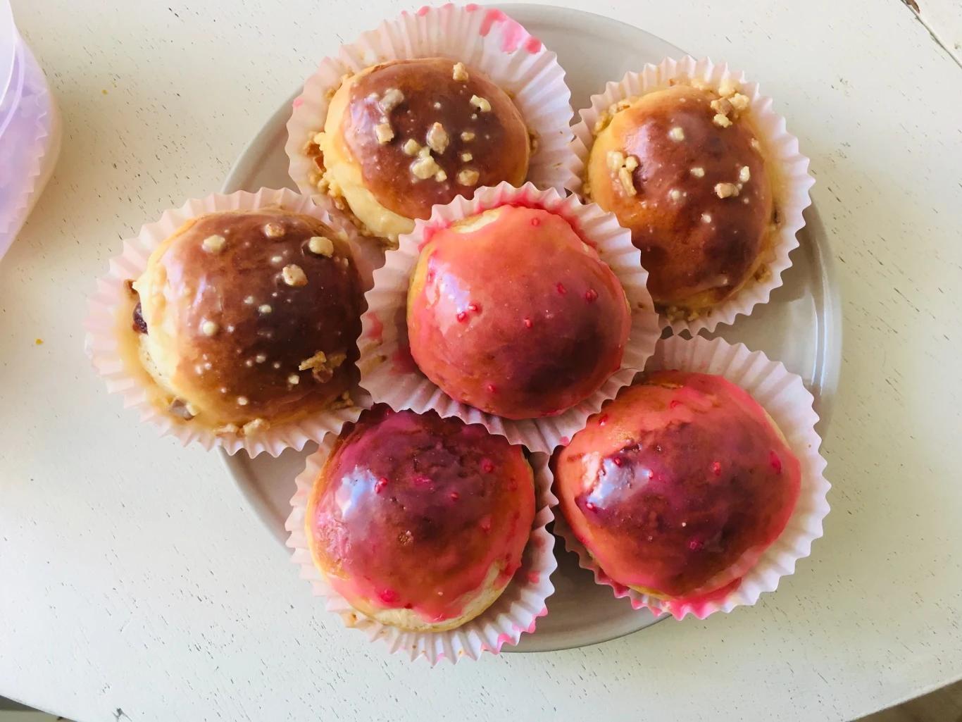 Sześć okrągłych, kolorowych bułeczek w papierowych foremkach, trzy z nich mają różową polewę, a trzy brązową z kawałkami orzechów, wszystko ułożone na talerzu.