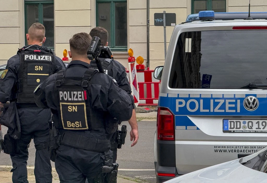 Niemcy zmagają się z falą przemocy ze strony gangów nieletnich (zdj. ilustracyjne) Trzech uzbrojonych niemieckich policjantów w kamizelkach kuloodpornych kieruje się w stronę budynku, obok radiowozu z napisem POLIZEI, na tle barier drogowych i miejskiej zabudowy.