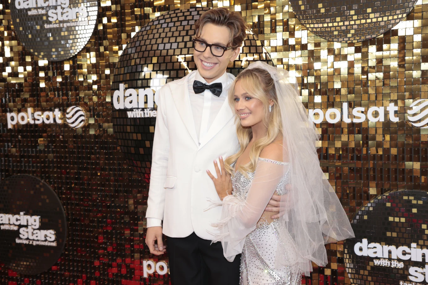 Para ubrana elegancko, mężczyzna w białym smokingu i kobieta w srebrnej sukni oraz welonie, pozują razem na tle błyszczącej ścianki z logotypami programu Dancing with the Stars oraz Polsat.