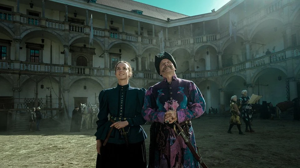 Dwie osoby w tradycyjnych, historycznych strojach stoją na dziedzińcu renesansowego zamku, w tle widoczne arkady i kilku ludzi w historycznych przebraniach.