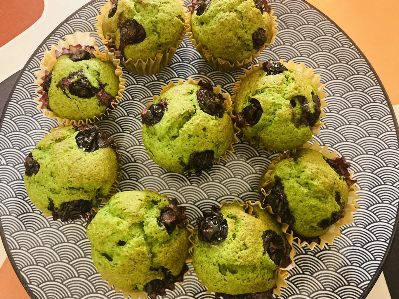 Siedem zielonych muffinów z widocznymi ciemnymi owocami, ustawionych okrężnie na dekoracyjnym talerzu z graficznym wzorem.