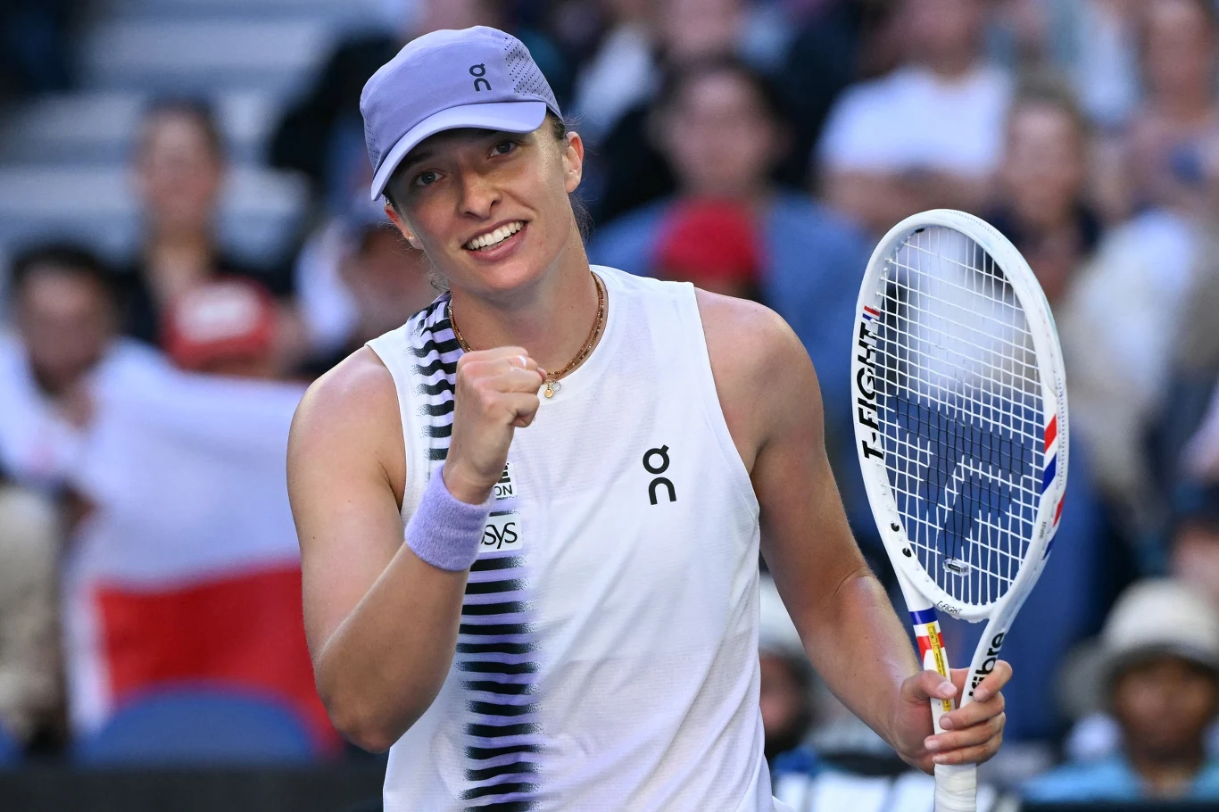 Tenisistka ubrana w sportowy strój i czapkę, trzyma rakietę tenisową, cieszy się z sukcesu na korcie, w tle widoczni kibice.