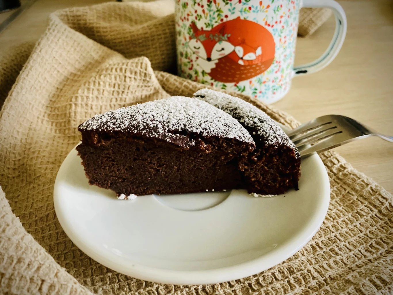 Kawałek ciasta czekoladowego posypanego cukrem pudrem leży na białym talerzyku, obok widelczyk na kremowym materiale, w tle kubek z kolorowym wzorem przedstawiającym rude, śpiące lisy.