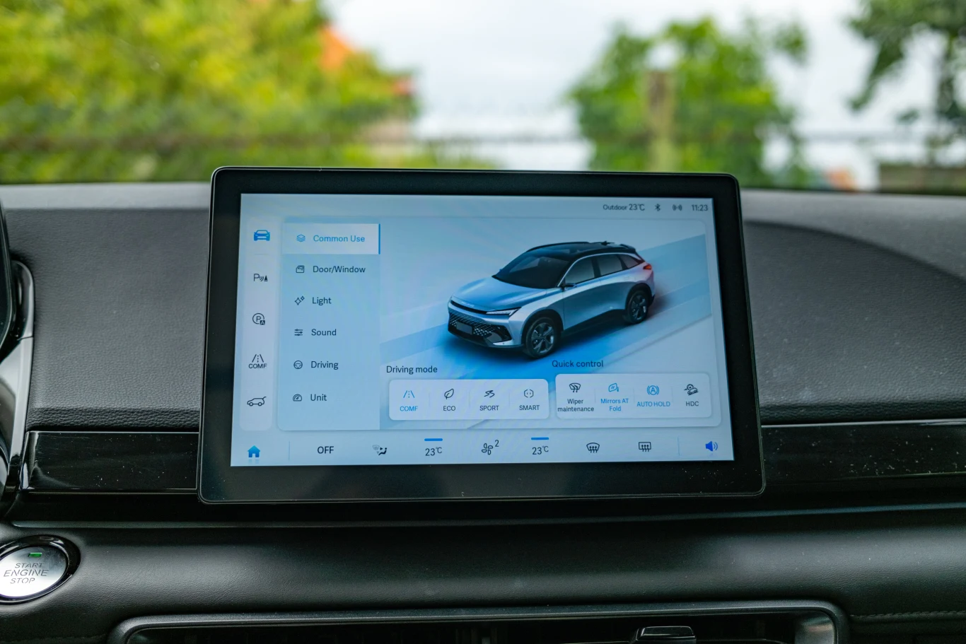Dotykowy ekran systemu multimedialnego w samochodzie wyświetlający ustawienia pojazdu i tryby jazdy, w tle widoczna deska rozdzielcza oraz rozmyte drzewa za szybą.