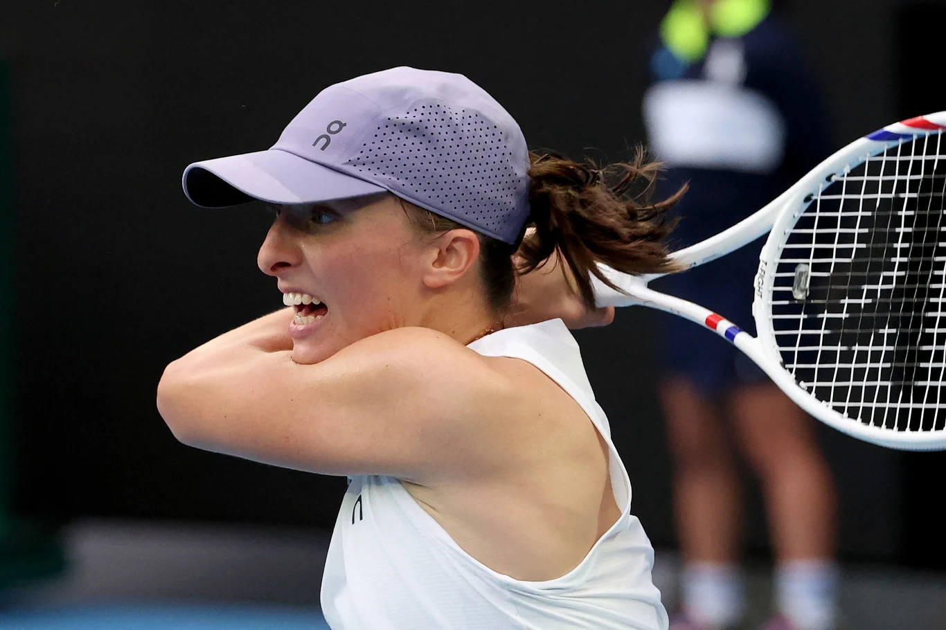 Iga Świątek na Australian Open 2026 Tenisistka w białej koszulce i fioletowej czapce podczas gry intensywnie macha rakietą, skupienie widoczne na jej twarzy i dynamicznie napięte mięśnie ramienia.