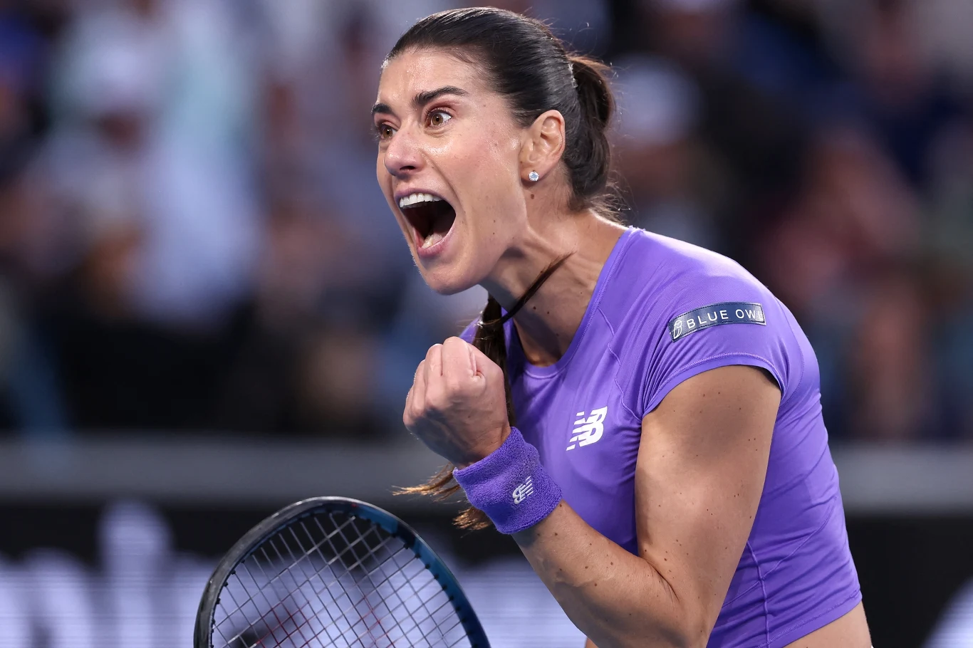 Tenisistka w fioletowej koszulce okazuje silne emocje po wygraniu punktu, ściska pięść i krzyczy z radości na korcie tenisowym. W tle widoczna rozmyta publiczność.