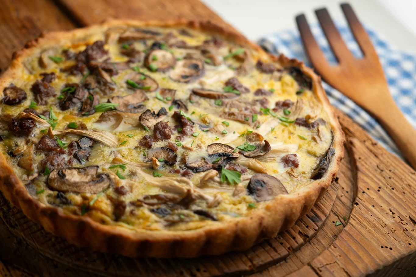 Tarta z delikatnym nadzieniem z grzybami, mięsem oraz ziołami, umieszczona na drewnianej desce, w tle widoczny drewniany widelec i fragment ściereczki w kratkę.