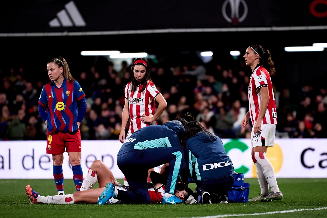 Kilka piłkarek na boisku, dwie z nich w strojach medycznych udzielają pomocy leżącej zawodniczce, zawodniczki z drużyn przeciwnych obserwują sytuację z widocznym zaniepokojeniem