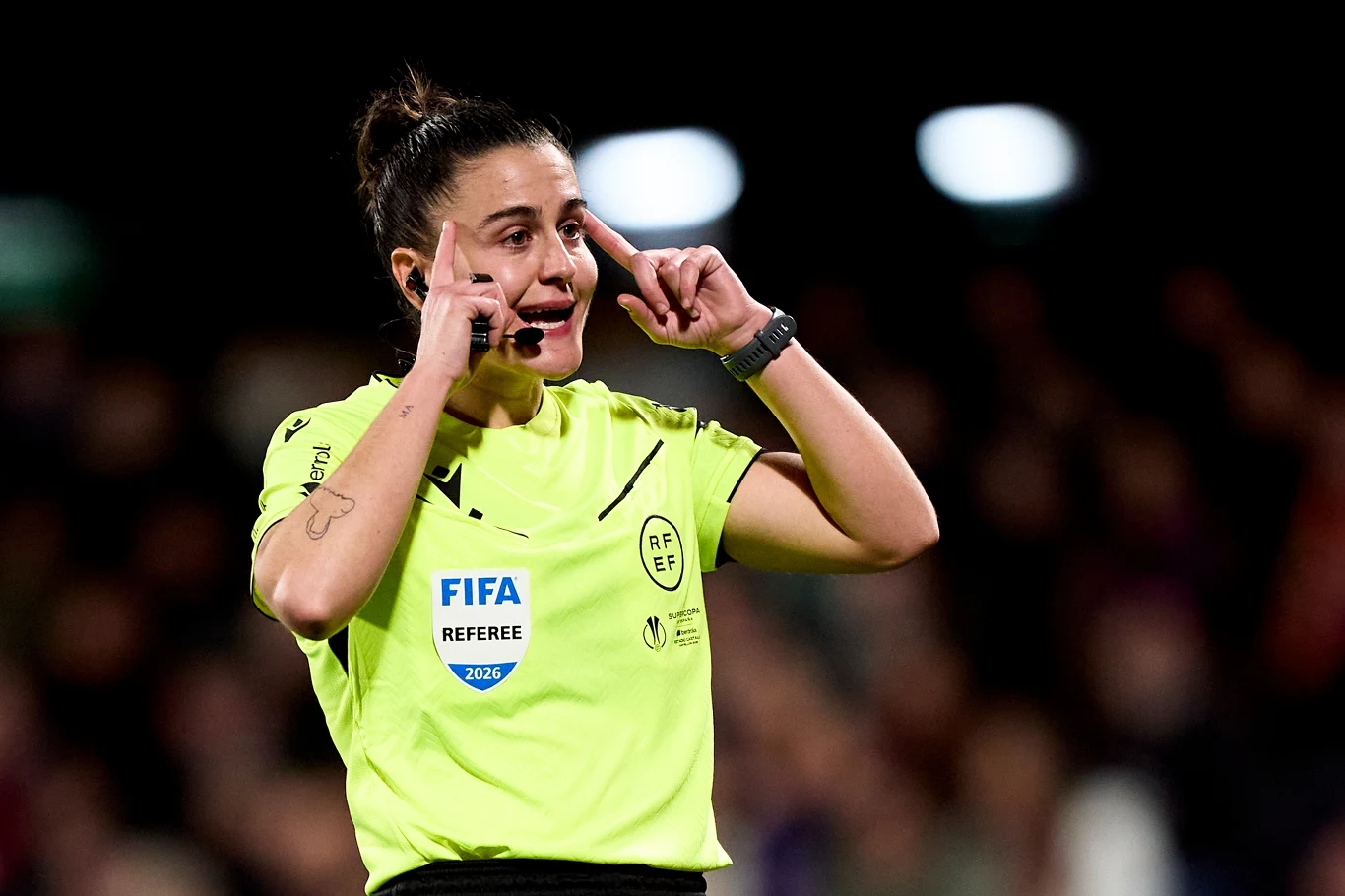 Sędzia piłkarski w jasnożółtej koszulce z odznaką FIFA wykonuje gest palcami przy skroniach, stojąc na tle zamazanego stadionu podczas meczu.