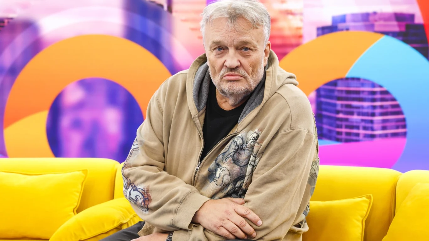 Krzysztof Cugowski Starszy mężczyzna z siwymi włosami i brodą siedzi na żółtej kanapie w nowoczesnym, kolorowym wnętrzu. Ubrany jest w beżową bluzę z nadrukiem, spogląda poważnie przed siebie. W tle widoczne są abstrakcyjne, kolorowe kształty i fragmenty zabudowy miejskiej.