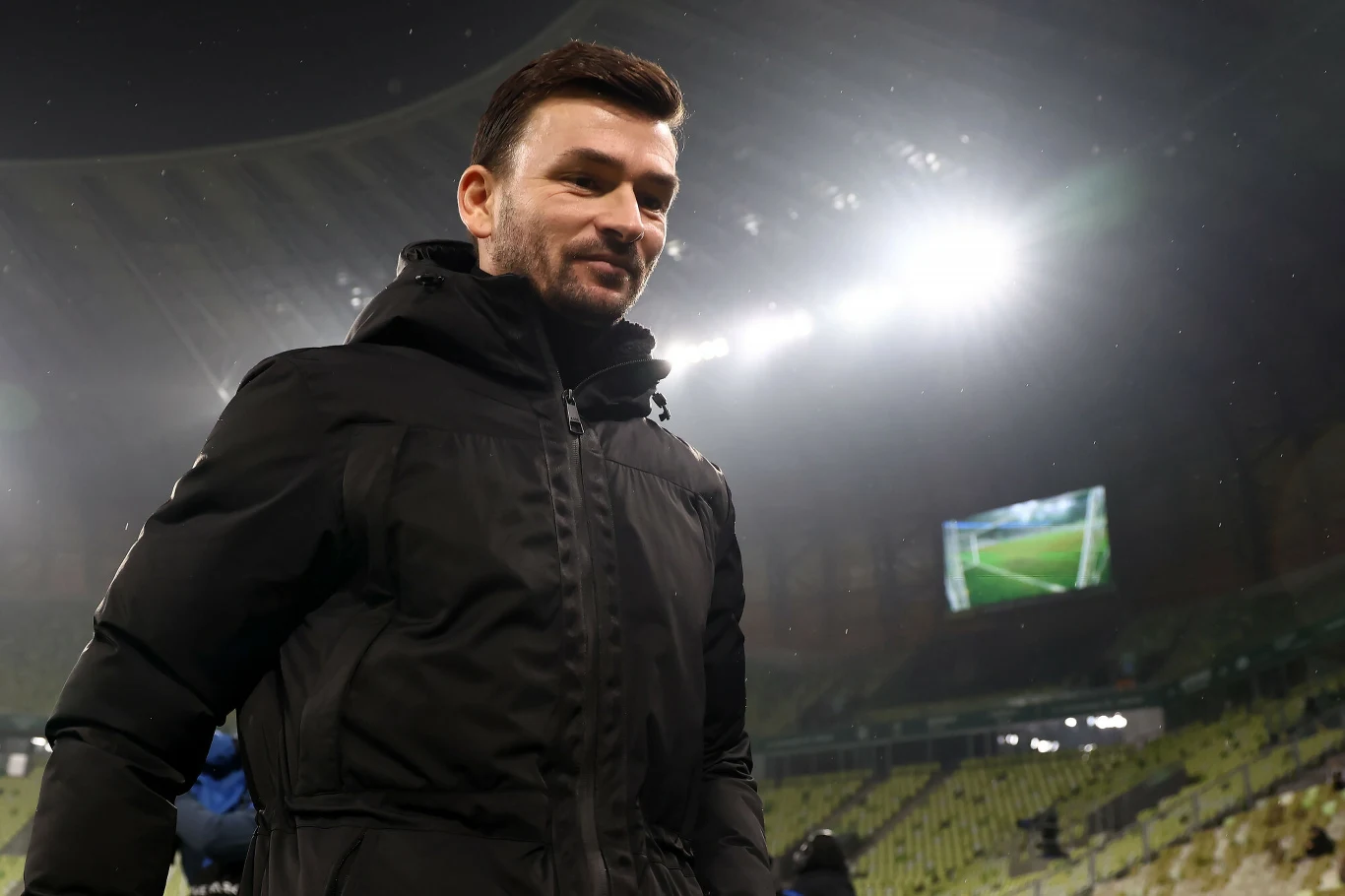 Mężczyzna w ciemnej kurtce stojący na tle pustego stadionu oświetlonego jasnymi reflektorami; w tle widoczny duży ekran z fragmentem boiska.