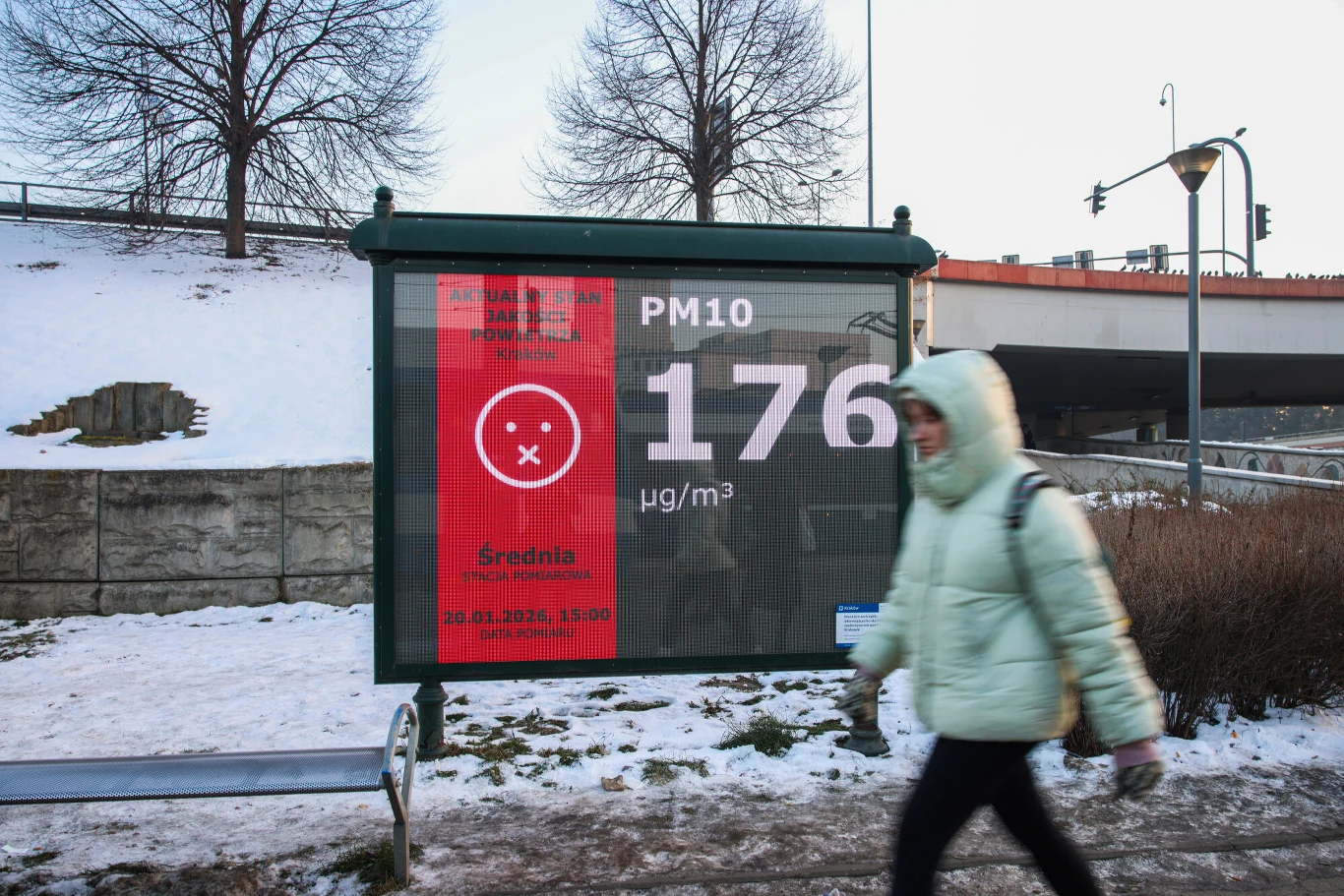 Przystanek autobusowy z dużym, elektronicznym wyświetlaczem informującym o poziomie pyłu PM10 wynoszącym 176 µg/m³. Przechodząca przed tablicą osoba ubrana w zimową kurtkę, w tle ośnieżony teren i fragment mostu.