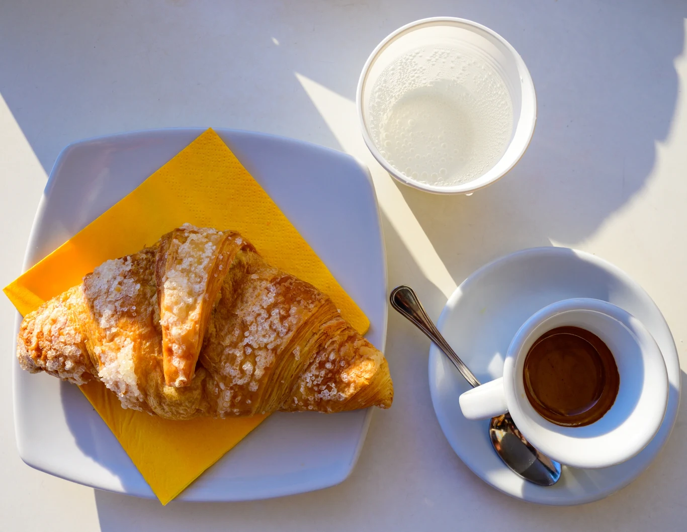 Espresso i cornetto to kultowe włoskie śniadanie Rogal z cukrem pudrem na białym talerzu z żółtą serwetką, filiżanka espresso na spodku z łyżeczką oraz plastikowy kubek z wodą ustawione na jasnym stole w naturalnym świetle.
