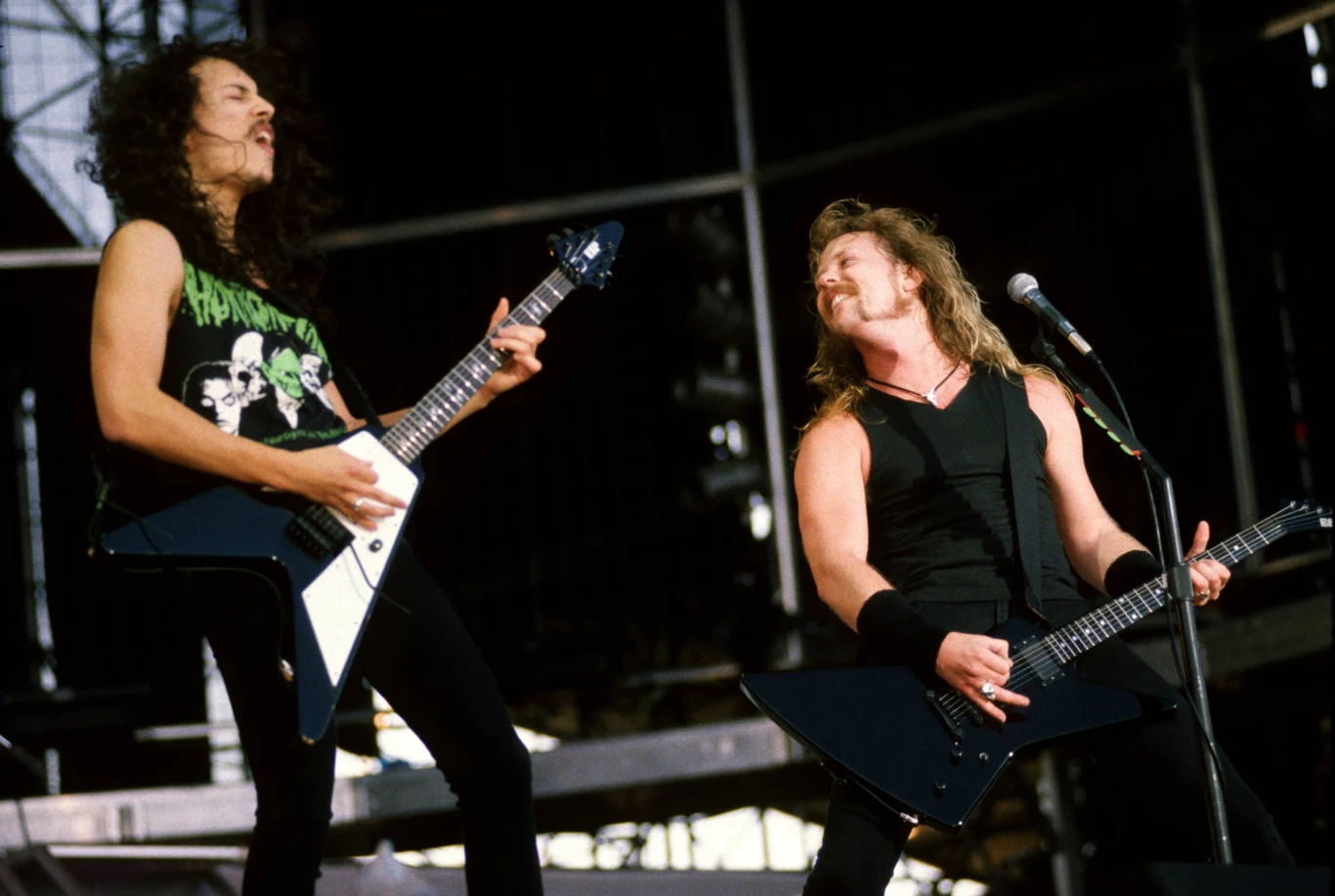Kirk Hammett i James Hetfield w 1991 roku Dwóch muzyków grających na gitarach elektrycznych na scenie koncertowej, wyrażających energię i zaangażowanie poprzez dynamiczny wyraz twarzy i postawy ciała. W tle widać elementy konstrukcji scenicznej i mikrofon.