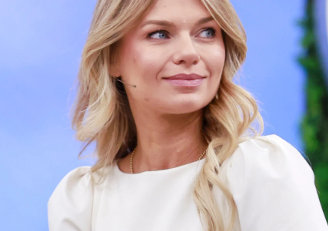 Emilia Komarnicka Blondynka o długich, falowanych włosach ubrana w jasną bluzkę, siedzi na tle niebieskiego tła z zielonym akcentem po prawej stronie, uśmiecha się subtelnie i patrzy w bok.