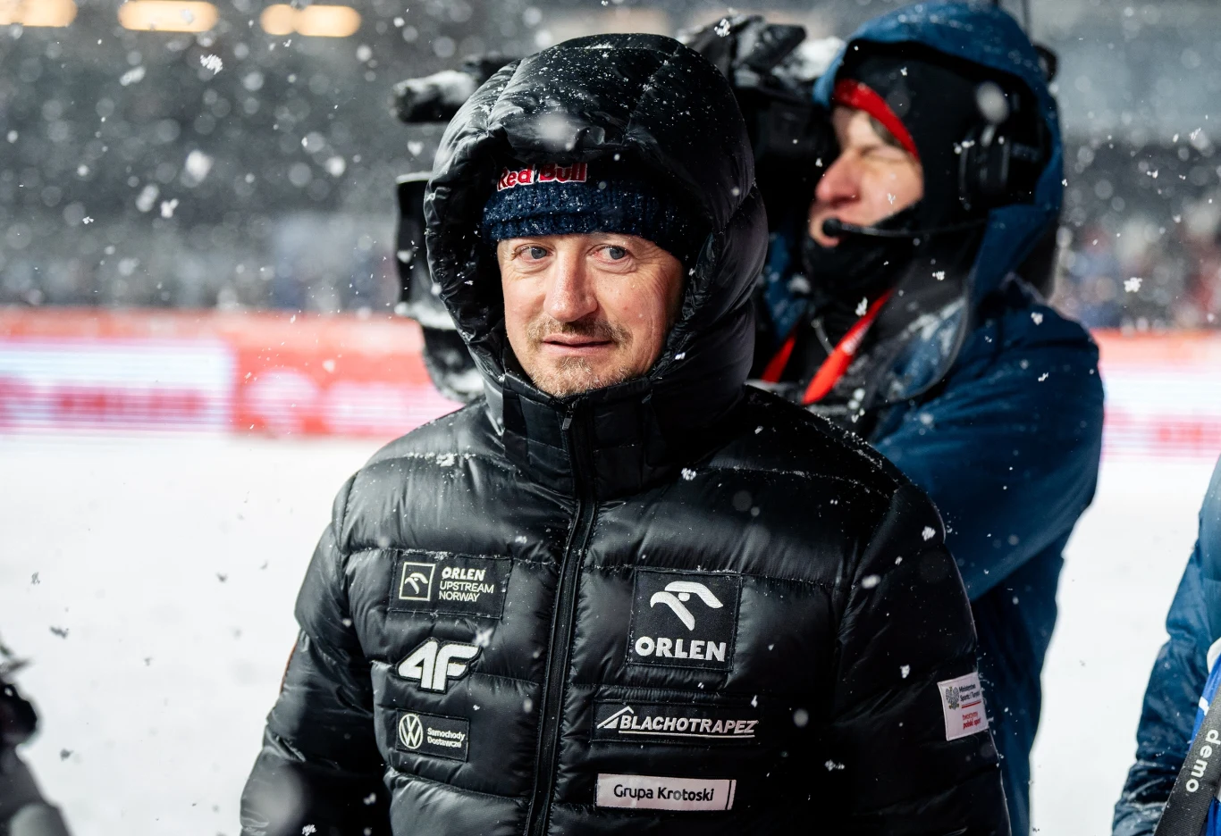 Mężczyzna ubrany w czarną zimową kurtkę z logotypami sponsorskimi stoi na śniegu, w tle widać inną osobę w ciepłym ubraniu, otoczenie sugeruje zimową imprezę sportową.