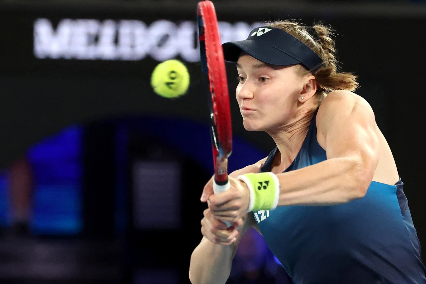 Zawodniczka tenisa uderza piłkę rakietą podczas meczu, intensywna koncentracja na twarzy, sportowe ubranie, opaska na głowie, dynamiczny ruch rakiety i piłki, w tle rozmazane napisy oraz ciemne otoczenie kortu.