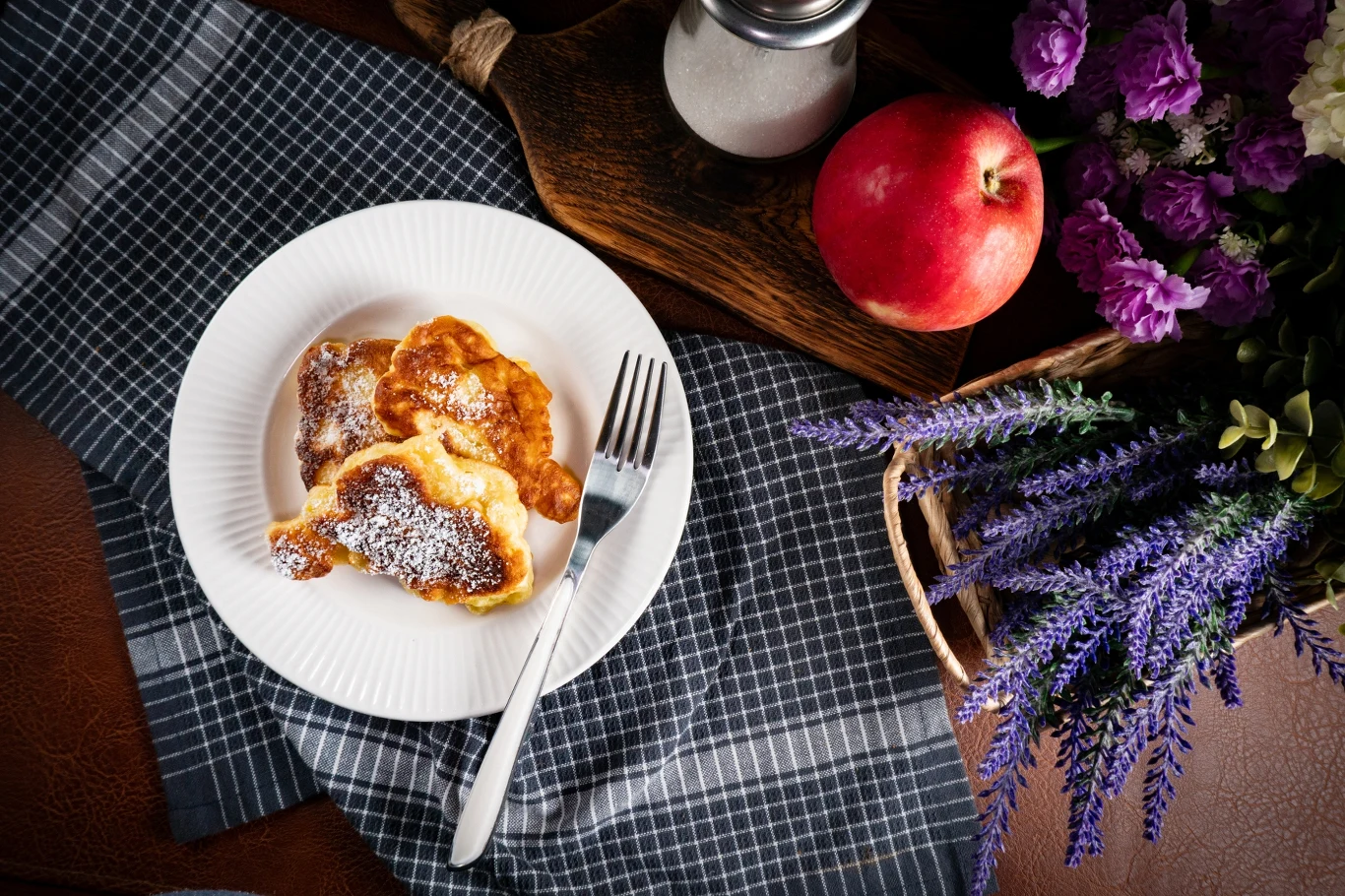 Porcja racuchów posypanych cukrem pudrem na białym talerzu, obok sztućców, położona na eleganckiej ściereczce, w tle jabłko, słoik z cukrem i kompozycja fioletowych kwiatów.
