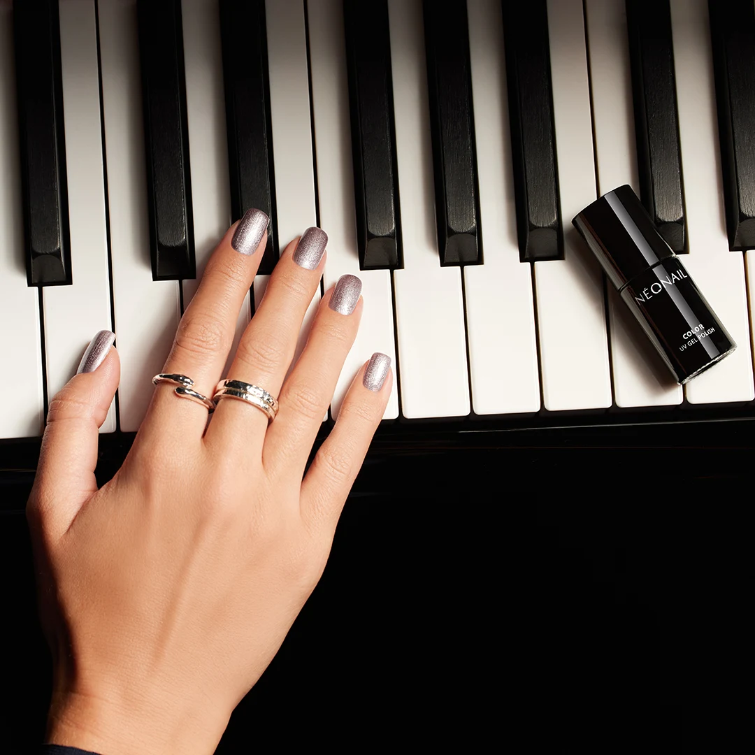 Dłoń z eleganckim manicure w srebrnym kolorze, ozdobiona pierścionkami, spoczywa na klawiaturze fortepianu. Obok palców widoczny jest lakier hybrydowy w czarnym opakowaniu.